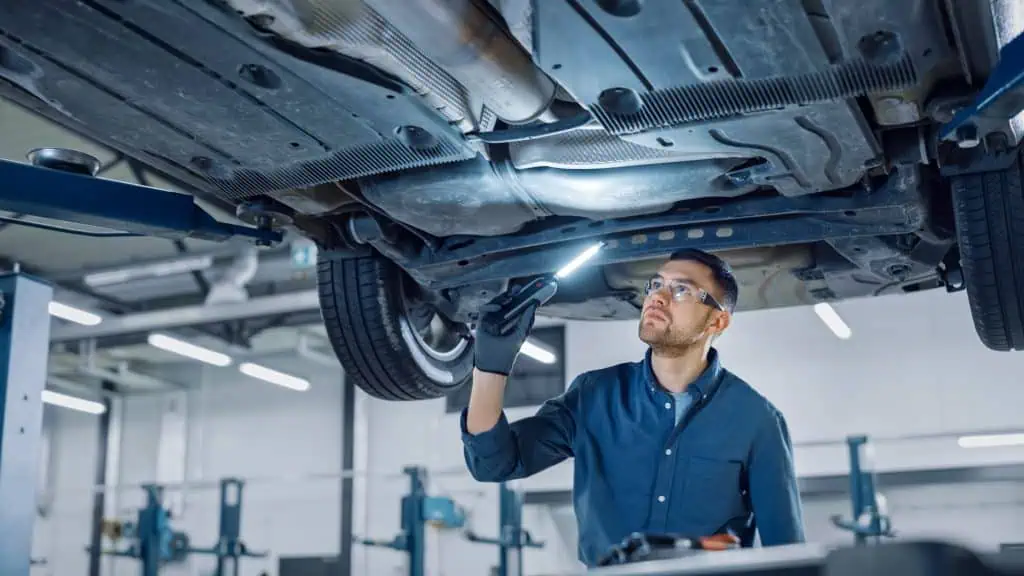 Handsome Professional Car Mechanic is Investigating Rust Under a Vehicle on a Lift in Service. Repairman is Using a LED lamp and Walks Towards. Specialist is Wearing Safety Glasses. Modern Workshop.